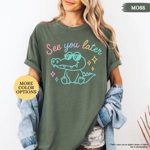 Puede incluir: Una camiseta verde musgo con la frase "See you later" y un cocodrilo de dibujos animados con gafas de sol. El cocodrilo est&aacute; delineado en azul y tiene un cuerpo azul claro. La camiseta tambi&eacute;n tiene el texto "MORE COLOR OPTIONS".
