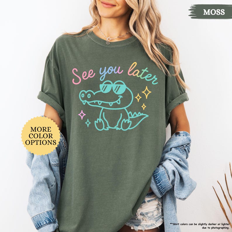 Puede incluir: Una camiseta verde musgo con la frase "See you later" y un cocodrilo de dibujos animados con gafas de sol. El cocodrilo est&aacute; delineado en azul y tiene un cuerpo azul claro. La camiseta tambi&eacute;n tiene el texto "MORE COLOR OPTIONS".