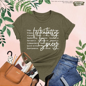K&ouml;nnte beinhalten: Olivgr&uuml;nes T-Shirt mit dem wei&szlig;en Schriftzug "Mentally Spicy". Das Shirt zeigt eine Liste von Adjektiven wie "Edgy", "Sassy" und "Fearless". Das Bild zeigt eine blaue Jeans und eine braune Tragetasche.