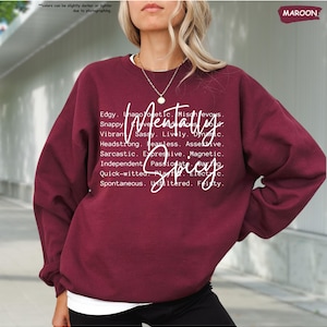 K&ouml;nnte beinhalten: Burgunderfarbenes Sweatshirt mit dem Schriftzug "Mentally Spicy" in wei&szlig;er Schrift. Das Design enth&auml;lt eine Liste von Adjektiven wie "Edgy", "Snappy" und "Sarcastic". Das Sweatshirt ist einfarbig und hat einen Rundhalsausschnitt.
