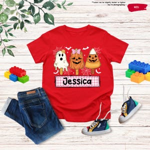 Może przedstawiać: Czerwony t-shirt z grafiką przedstawiającą trzy lody na patyku w stylu Halloween: duszka, dynię i cukierek kukurydziany. Poniżej znajduje się r&oacute;żowo-biały prostokąt w kratkę z imieniem Jessica napisanym czarną, stylizowaną czcionką.