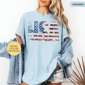 May include: Light blue t-shirt with a distressed American flag design. The flag graphic features red and white stripes and a blue field of stars, with the word "USA" incorporated into the design. The shirt has rolled-up sleeves and a casual fit.