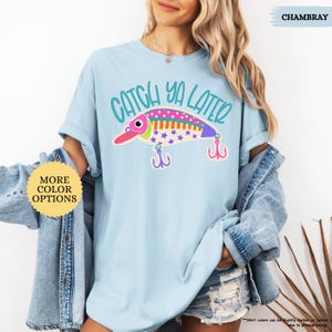 以下が含まれることがあります： 水色のTシャツで、釣り用ルアーのグラフィックの上に「CATCH YA LATER」という文字がティール色でプリントされています。ルアーはピンク、オレンジ、青のストライプ、水玉模様、青い星で装飾されており、下に2つの紫色のトリプルフックがぶら下がっています。