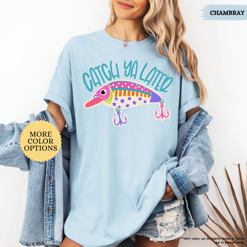 以下が含まれることがあります： 水色のTシャツで、釣り用ルアーのグラフィックの上に「CATCH YA LATER」という文字がティール色でプリントされています。ルアーはピンク、オレンジ、青のストライプ、水玉模様、青い星で装飾されており、下に2つの紫色のトリプルフックがぶら下がっています。