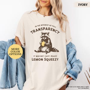 May include: A light beige t-shirt with a raccoon graphic holding a lemon. The text on the shirt reads "In the interest of full transparency, it was not easy peasy lemon squeezy".