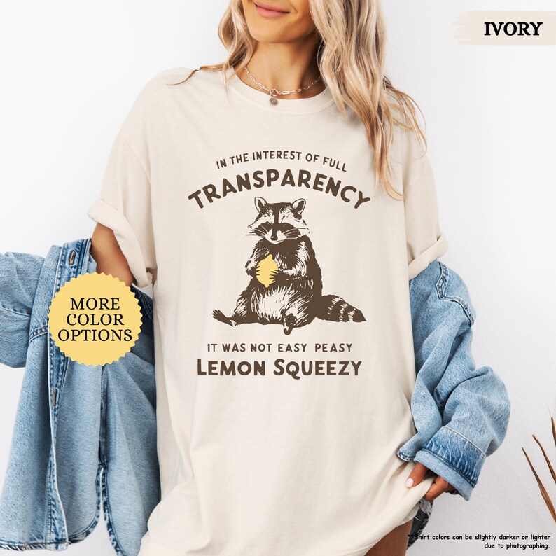 May include: A light beige t-shirt with a raccoon graphic holding a lemon. The text on the shirt reads "In the interest of full transparency, it was not easy peasy lemon squeezy".