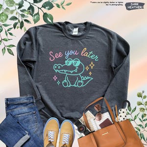 May include: Dark heather gray sweatshirt with a cartoon alligator wearing sunglasses and the phrase "See you later" in colorful text. The design includes sparkling stars. The sweatshirt is displayed with jeans, shoes, and a handbag.