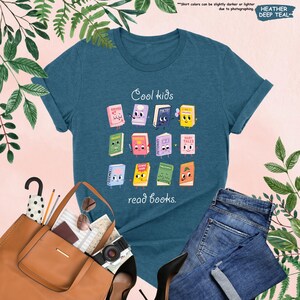 Puede incluir: Una camiseta de color verde azulado oscuro con el texto "Cool kids read books". La camiseta presenta un gr&aacute;fico de libros de dibujos animados con caras. Tambi&eacute;n se ve un bolso tote de cuero marr&oacute;n, gafas de sol y jeans.