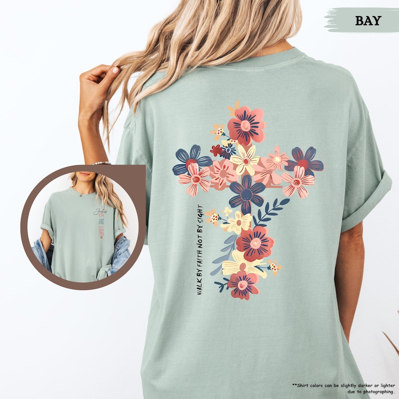May include: A sage green t-shirt features a floral cross design with pink, peach, and blue flowers.  The text 'Walk by faith not by sight' is vertically oriented along the side of the cross. The shirt is a relaxed fit, short-sleeved tee.  The back of the shirt is shown in the main image.