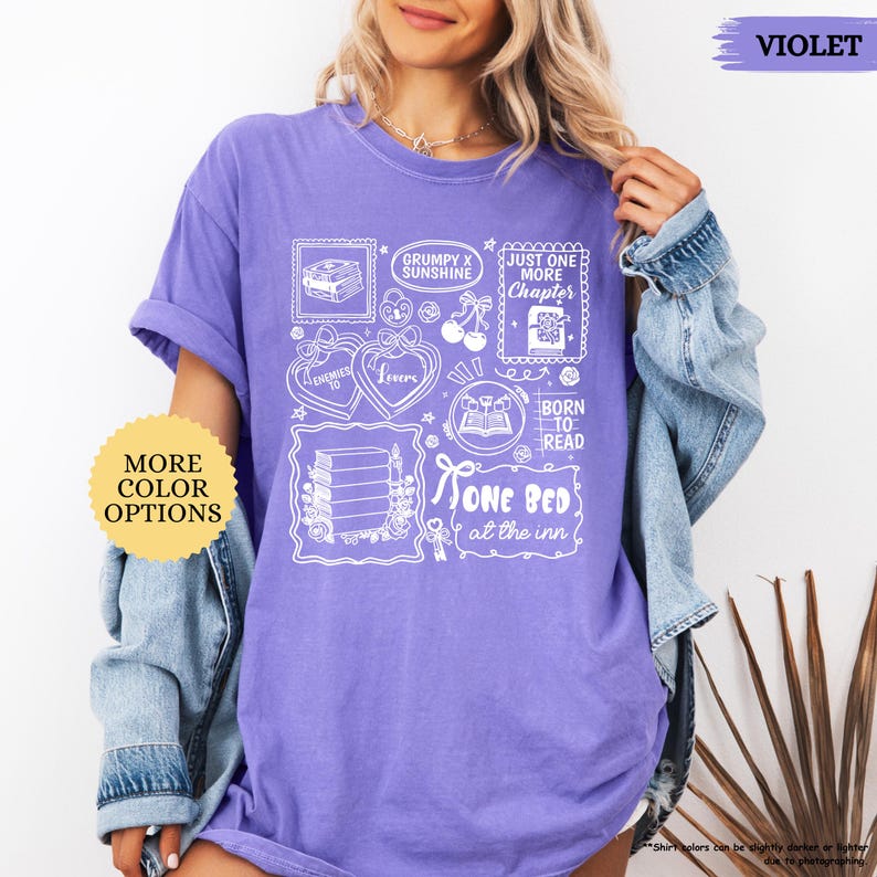 Puede incluir: Una camiseta de color violeta con dise&ntilde;os gr&aacute;ficos blancos, incluyendo pilas de libros, corazones y el texto "Just One More Chapter". La camiseta tambi&eacute;n presenta los textos "Grumpy x Sunshine" y "Born to Read".