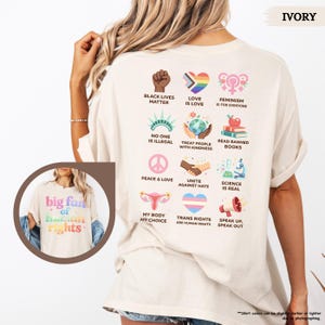 May include: Ivory-colored t-shirt with the text "big fan of human rights" and various graphic symbols representing social justice and equality. The back of the shirt features additional icons and phrases like "Black Lives Matter" and "Love is Love."