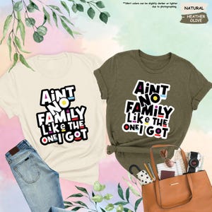 May include: Two t-shirts, one cream and one olive green, with the text "Ain't No Family Like The One I Got" in a colorful, bold font. The cream shirt is paired with blue jeans. The olive shirt is styled with a brown tote bag and accessories.