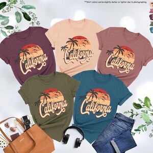 May include: Five t-shirts with a sunset and palm tree graphic and the text "California" in a retro font. The shirts are in various colors including heather peach, heather mauve, heather maroon, heather olive, and heather deep teal.