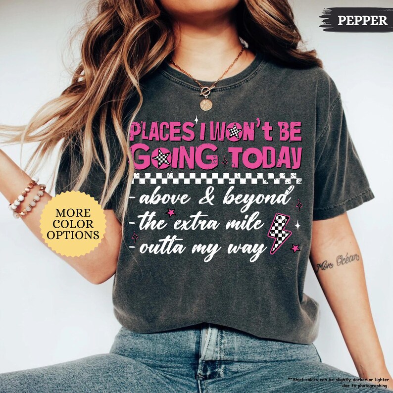 May include: Dark gray t-shirt with the text "PLACES I WON'T BE GOING TODAY" in pink and white. The shirt features a checkered flag and lightning bolt graphic. A button on the left says "MORE COLOR OPTIONS".