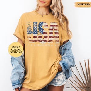 May include: Mustard-colored t-shirt with a distressed USA flag design in red, white, and blue. The text "SINCE 1776" is below the flag. The shirt has rolled-up sleeves and is paired with a denim jacket and shorts. The words "MORE COLOR OPTIONS" are also visible.