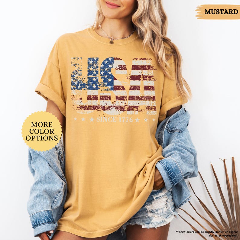 May include: Mustard-colored t-shirt with a distressed USA flag design in red, white, and blue. The text "SINCE 1776" is below the flag. The shirt has rolled-up sleeves and is paired with a denim jacket and shorts. The words "MORE COLOR OPTIONS" are also visible.