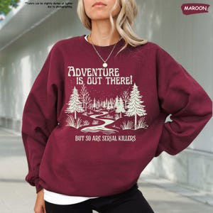 May include: Maroon sweatshirt with the text "ADVENTURE IS OUT THERE! BUT SO ARE SERIAL KILLERS" in white. The graphic features a winding path through a forest with trees and a starry sky. The sweatshirt is a long-sleeved crew neck.