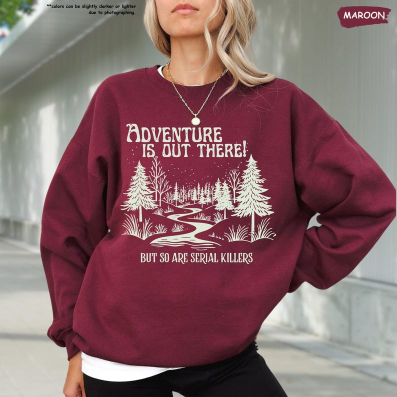 May include: Maroon sweatshirt with the text "ADVENTURE IS OUT THERE! BUT SO ARE SERIAL KILLERS" in white. The graphic features a winding path through a forest with trees and a starry sky. The sweatshirt is a long-sleeved crew neck.