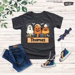 Może przedstawiać: Ciemnoszary melanżowy t-shirt z grafiką przedstawiającą trzy lody na patyku w stylu Halloween: duszka, dynię w czapce oraz cukierek kukurydziany. Pod lodami znajduje się pomarańczowo-biały prostokąt w kratkę z napisem Thomas.