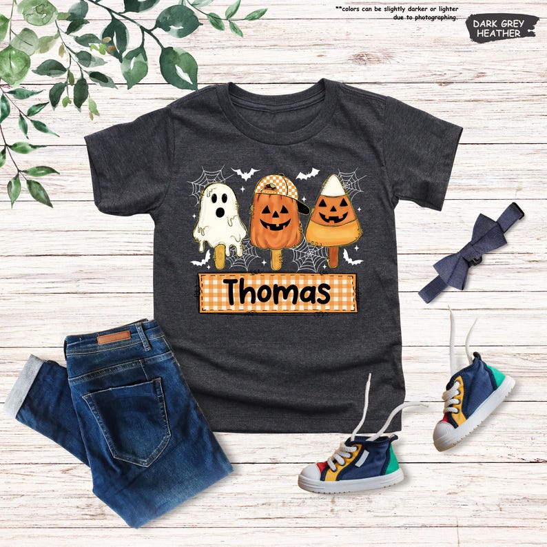 Może przedstawiać: Ciemnoszary melanżowy t-shirt z grafiką przedstawiającą trzy lody na patyku w stylu Halloween: duszka, dynię w czapce oraz cukierek kukurydziany. Pod lodami znajduje się pomarańczowo-biały prostokąt w kratkę z napisem Thomas.