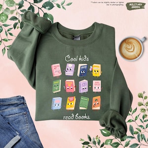 Puede incluir: Sudadera verde militar con el texto "Cool kids read books." El dise&ntilde;o presenta libros de dibujos animados con caras. La sudadera se combina con vaqueros azules y una taza de caf&eacute;. El color puede variar ligeramente.