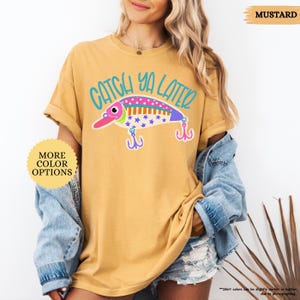以下が含まれることがあります： マスタードイエローのTシャツ。ティール色の文字で「CATCH YA LATER」と書かれ、その下にカラフルな釣り用ルアーのグラフィックが描かれています。ルアーはピンク、白、紫で、青い星模様と2つのフックが付いています。「MORE COLOR OPTIONS」と書かれた黄色い円形のマークが表示されています。