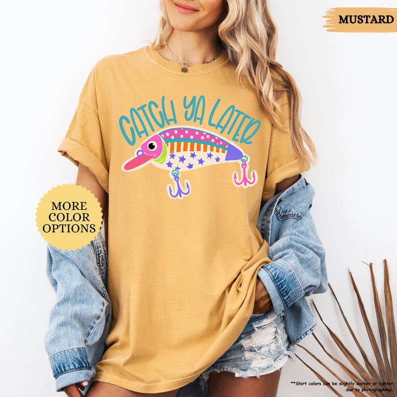 以下が含まれることがあります： マスタードイエローのTシャツ。ティール色の文字で「CATCH YA LATER」と書かれ、その下にカラフルな釣り用ルアーのグラフィックが描かれています。ルアーはピンク、白、紫で、青い星模様と2つのフックが付いています。「MORE COLOR OPTIONS」と書かれた黄色い円形のマークが表示されています。