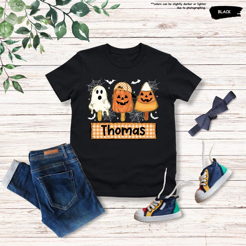 Może przedstawiać: Czarna koszulka z grafiką przedstawiającą trzy lody na patyku w stylu Halloween: białego ducha, pomarańczową dynię w czapce oraz cukierek kukurydziany, wszystkie z twarzami dyni. Pod grafiką znajduje się pomarańczowy prostokąt w kratkę z imieniem Thomas.