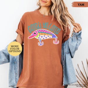 以下が含まれることがあります： 錆色の半袖Tシャツで、カラフルな釣り用ルアーのグラフィックとティール色の「CATCH YA LATER」という文字がプリントされています。ルアーのデザインには、ピンク、オレンジ、青、白の星やドットの模様があり、2つのフックが付いています。