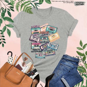 May include: Athletic heather gray t-shirt featuring a colorful graphic of cassette tapes with song titles like "Jesus Anthems" and "Reckless Love." The shirt is paired with blue jeans, a brown tote bag, and black heels. The text "He Knows My Name" is visible.