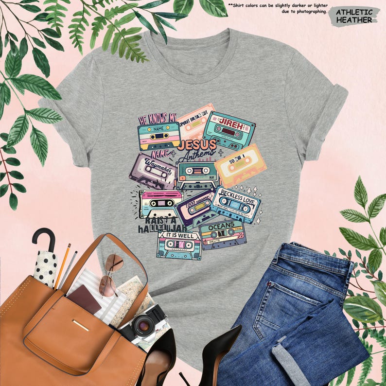 May include: Athletic heather gray t-shirt featuring a colorful graphic of cassette tapes with song titles like "Jesus Anthems" and "Reckless Love." The shirt is paired with blue jeans, a brown tote bag, and black heels. The text "He Knows My Name" is visible.