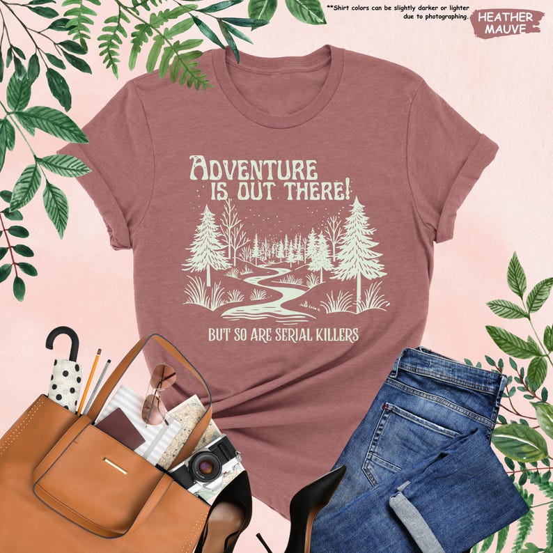 May include: Heather mauve t-shirt with the text "ADVENTURE IS OUT THERE! BUT SO ARE SERIAL KILLERS" and a forest graphic. The shirt is paired with blue jeans, a brown purse, and black heels.