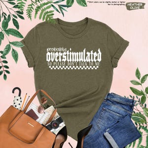 May include: A green t-shirt with white text that reads "probably overstimulated so please don't talk to me". The text is in a bold, gothic font with a checkered pattern underneath.