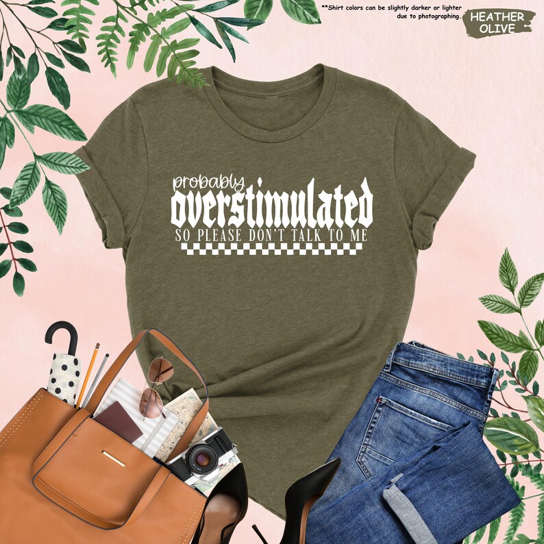 May include: A green t-shirt with white text that reads "probably overstimulated so please don't talk to me". The text is in a bold, gothic font with a checkered pattern underneath.
