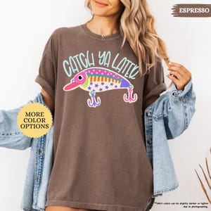 以下が含まれることがあります： エスプレッソカラーの半袖Tシャツ。カラフルな釣り用ルアーのグラフィックと、水色で「CATCH YA LATER」という文字がプリントされています。ルアーのデザインには、ピンク、オレンジ、白のセクションがあり、青い星とドットの模様が施されています。