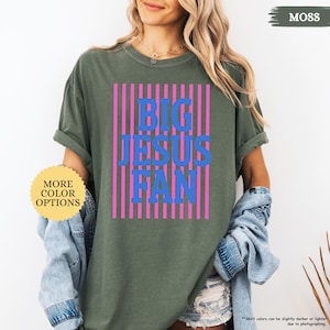 May include: A moss green t-shirt with the text "BIG JESUS FAN" in blue, set against pink vertical stripes. The shirt has rolled-up sleeves and a casual fit. A button with the text "MORE COLOR OPTIONS" is visible.