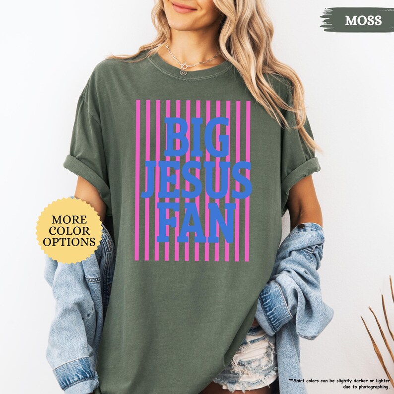 May include: A moss green t-shirt with the text "BIG JESUS FAN" in blue, set against pink vertical stripes. The shirt has rolled-up sleeves and a casual fit. A button with the text "MORE COLOR OPTIONS" is visible.
