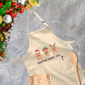 Op de afbeelding: Een beige katoenen schort met een zak en verstelbare banden. Het heeft een afbeelding van drie gingerbreadmensen met kerstpetten en de tekst "Christmas Baking Crew".