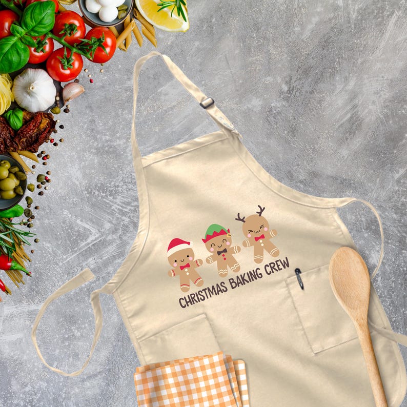 Op de afbeelding: Een beige katoenen schort met een zak en verstelbare banden. Het heeft een afbeelding van drie gingerbreadmensen met kerstpetten en de tekst "Christmas Baking Crew".