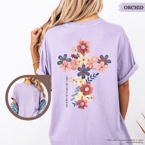 May include: A lavender t-shirt with a floral cross design and the text "Walk by faith not by sight".