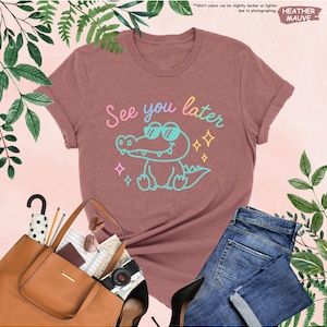 May include: Mauve t-shirt with a cartoon alligator wearing sunglasses and the phrase "See you later" in pastel colors. The shirt is paired with blue jeans, a brown purse, and black heels. The text "HEATHER MAUVE" is in the upper right corner.