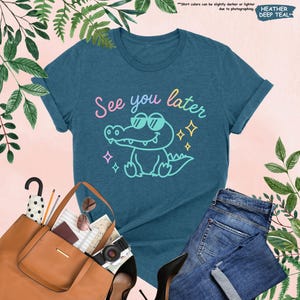 May include: A teal t-shirt with the phrase "See you later" and a cartoon alligator wearing sunglasses. The graphic is in pastel colors. The shirt is paired with blue jeans, a brown tote bag, and black heels.