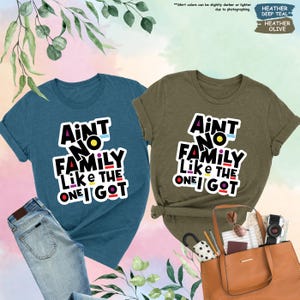 May include: Two t-shirts, one deep teal and one heather olive, each with the text "Ain't No Family Like The One I Got" in a colorful, bold font. The teal shirt is paired with jeans, while the olive shirt is next to a brown tote bag.