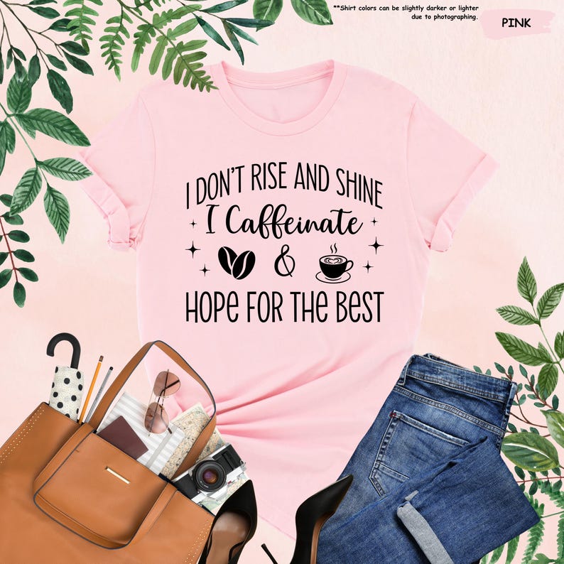 May include: Pink t-shirt with the text "I DON'T RISE AND SHINE I Caffeinate & HOPE FOR THE BEST". The shirt is styled with a brown purse, sunglasses, a camera, black heels, and blue jeans. Green leaves are in the background.