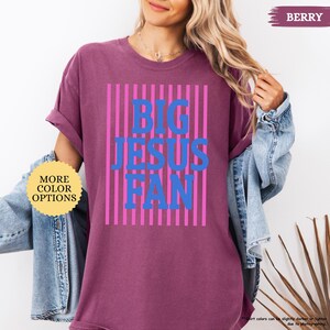 May include: Berry-colored t-shirt with the text "BIG JESUS FAN" in blue, set against pink vertical stripes. The shirt has rolled-up sleeves and a round neckline. A button on the left side says "MORE COLOR OPTIONS".