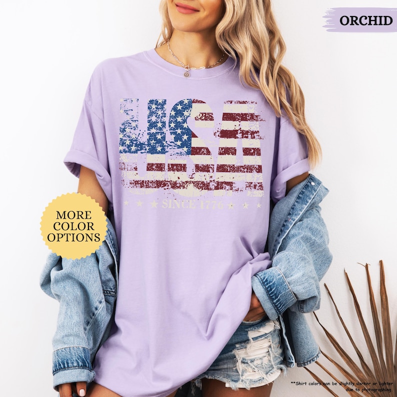 May include: Orchid-colored t-shirt with a distressed USA flag design. The flag graphic features red and white stripes, blue stars, and the text "USA SINCE 1776". The shirt has short sleeves and a relaxed fit. "More color options" is also visible.