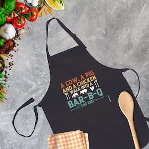 May include: Black apron with a white graphic that reads "A cow, a pig and a chicken walk into a bar-b-q - The end". The graphic is in a retro style with a sunset color scheme.