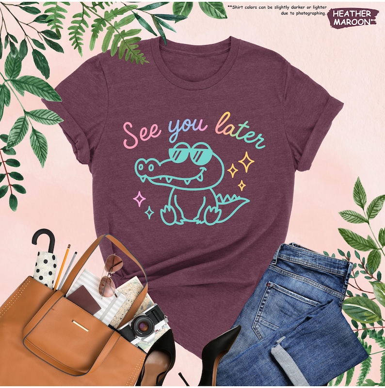 May include: Maroon t-shirt with the text "See you later" and a cartoon alligator wearing sunglasses. The design is in shades of blue, pink, and yellow. The shirt is paired with blue jeans, a brown tote bag, and black heels.