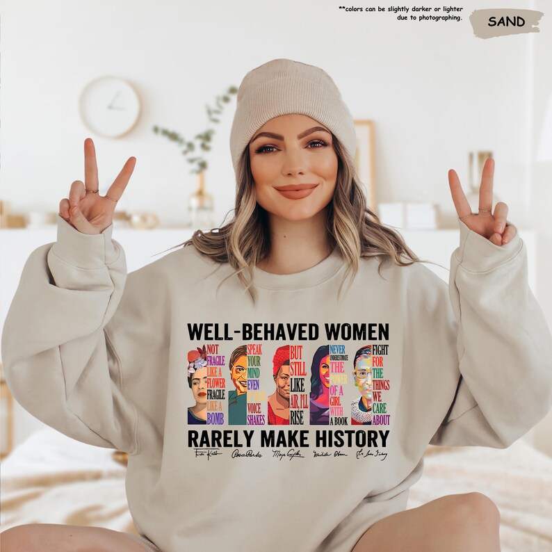 May include: A light beige sweatshirt with the text "Well-Behaved Women Rarely Make History" and illustrations of famous women, including Frida Kahlo,  with quotes from each woman. The text includes the names of the women: Frida Kahlo,  Alice Walker, Maya Angelou, Michelle Obama, and Ruth Bader Ginsburg.