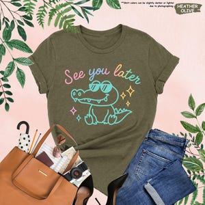 May include: Heather olive green t-shirt with the phrase "See you later" and a cartoon alligator wearing sunglasses. The shirt is paired with blue jeans, a brown tote bag, and black heels.